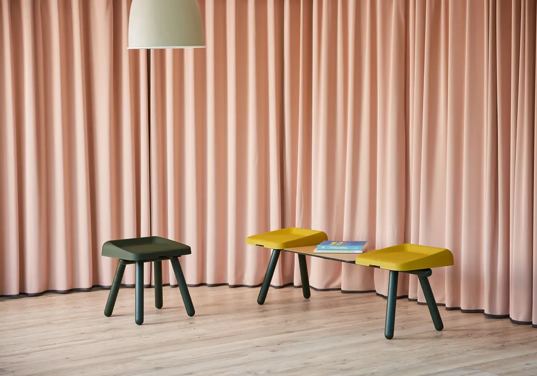 Taburete Slim verde y banco Slim amarillo con tablero de madera frente a una cortina rosada
